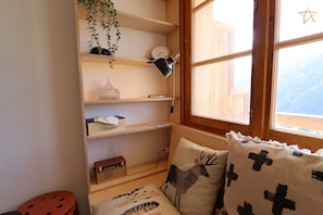 A cozy room with a wooden shelf unit, a radio, a window with a view, and a bed with patterned pillows.