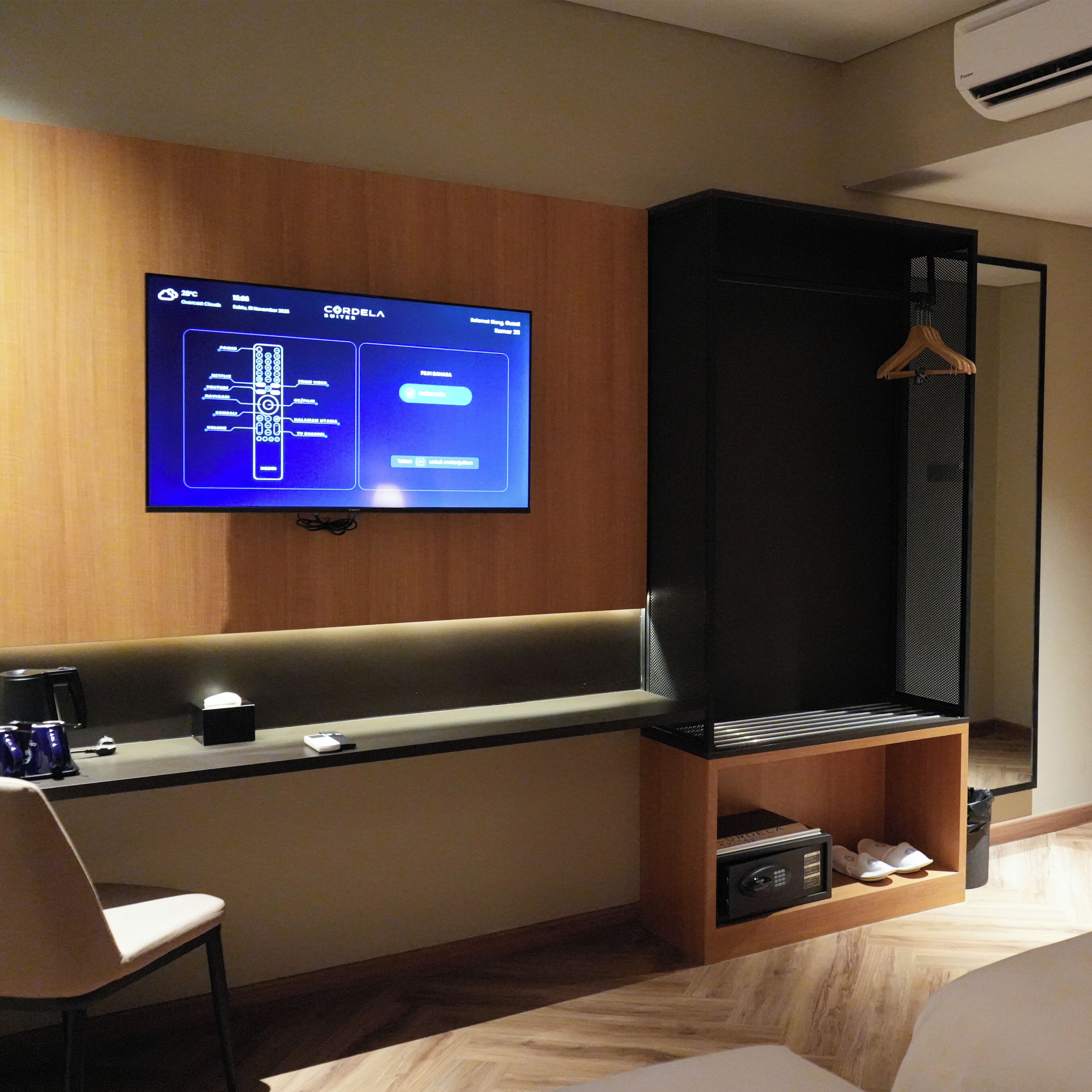 A modern hotel room with a flat-screen TV mounted on the wall, a wooden bench, a desk with a coffee machine, a coat rack, and a shoe storage area.