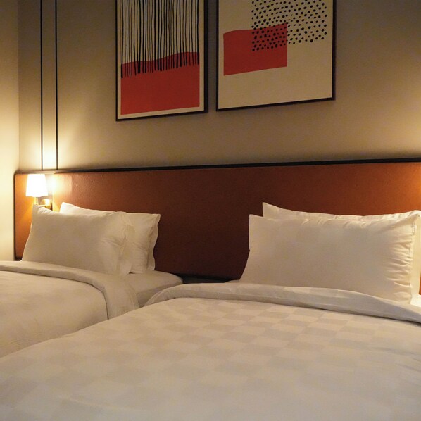 A hotel room with two beds, a nightstand with a lamp, and two abstract wall art pieces.