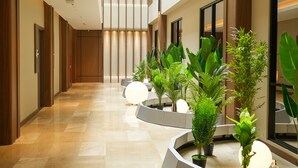 Un couloir d’hôtel moderne, avec un sol en marbre poli, des lambris en bois sur les murs et une série de plantes en pot disposées le long du passage.