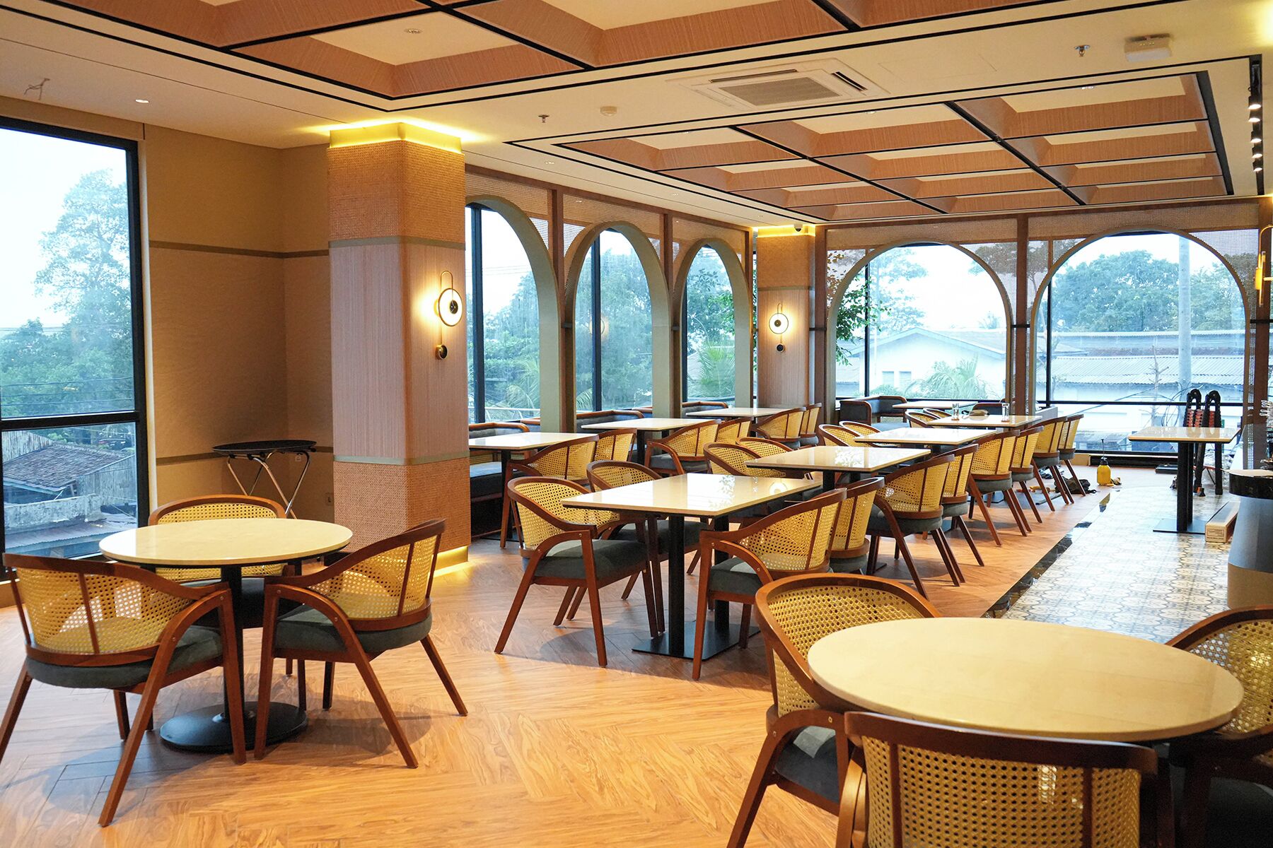 Un restaurant avec des tables rondes et des chaises en osier, de grandes fenêtres surmontées d’arcs, et une vue sur la verdure environnante.