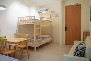 Huoneessa on kerrossängyt, puinen pöytä tuoleineen, sohva ja ovi.