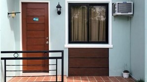 A front view of a building with a wooden door, a window with curtains, an air conditioning unit, and a small potted plant. - Sunshine Stay Panglao (Panglao)