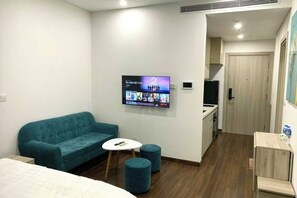 A hotel room with a teal sofa, a small white table, a TV mounted on the wall, a wooden floor, and a kitchenette area. - LISA HOUSE - MASTERI WEST HEIGHTS - VINHOMES SMART CITY (Hanoi)