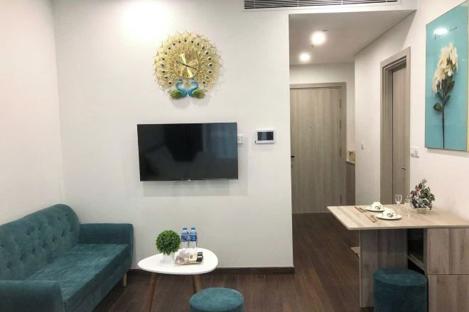 Un salon moderne avec un canapé de couleur turquoise, un téléviseur à écran plat, une horloge murale décorative, une petite table ronde agrémentée d’une plante et de bouteilles d’eau, et un coin repas avec une table et des chaises en bois.