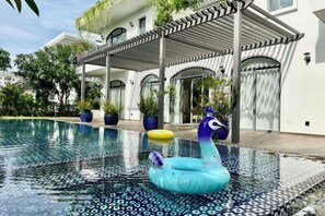 Une piscine agrémentée d’un cygne flottant, un patio couvert et un bâtiment au style architectural moderne.