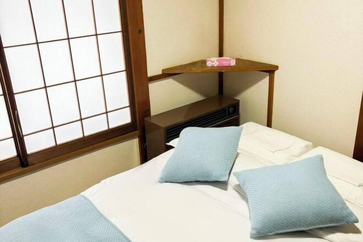 Quarto com cama, almofadas, uma pequena estante e janela com padrão quadriculado.