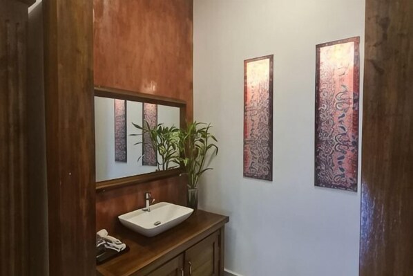 A bathroom with a wooden vanity, a white sink, a mirror, and two patterned wall decorations. - Villa Romduol (Kampot)
