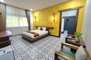 A hotel room with a bed, a desk, a chair, a small table with a plant, and a patterned floor. - Villa Romduol (Kampot)