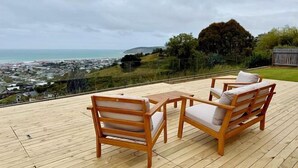 Une terrasse en bois avec deux chaises en bois et une petite table, d’où l’on a vue sur une ville côtière et des collines.