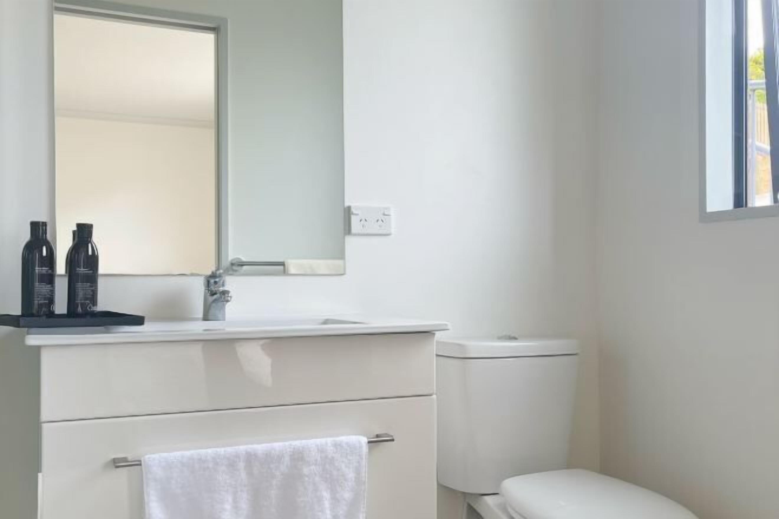 Een moderne badkamer met een wit wastafel, spiegel en toilet. Er staan twee flessen op de wastafel.