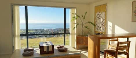 Una habitación con un ventanal que ofrece vistas al mar y al paisaje urbano. Hay una mesa de madera con una silla, un pequeño sofá con cojines y un aparador con una planta.