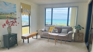 Un salon comprenant un canapé, deux petites tables, un banc, un vase décoratif et une grande fenêtre donnant sur la mer.