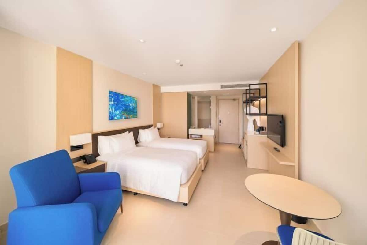 A hotel room with a bed, two bedside tables, a TV, a small table, and a blue chair.