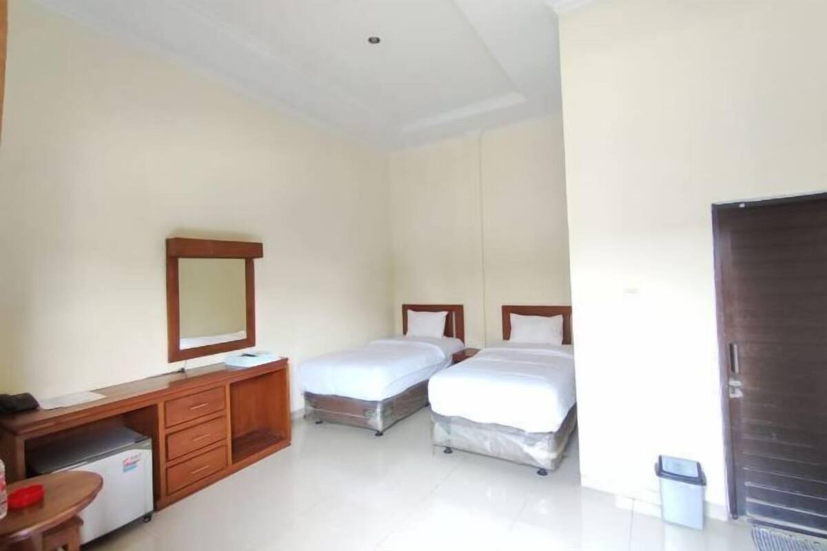A hotel room with two single beds, a wooden desk with a mirror, a small table, and a trash bin.