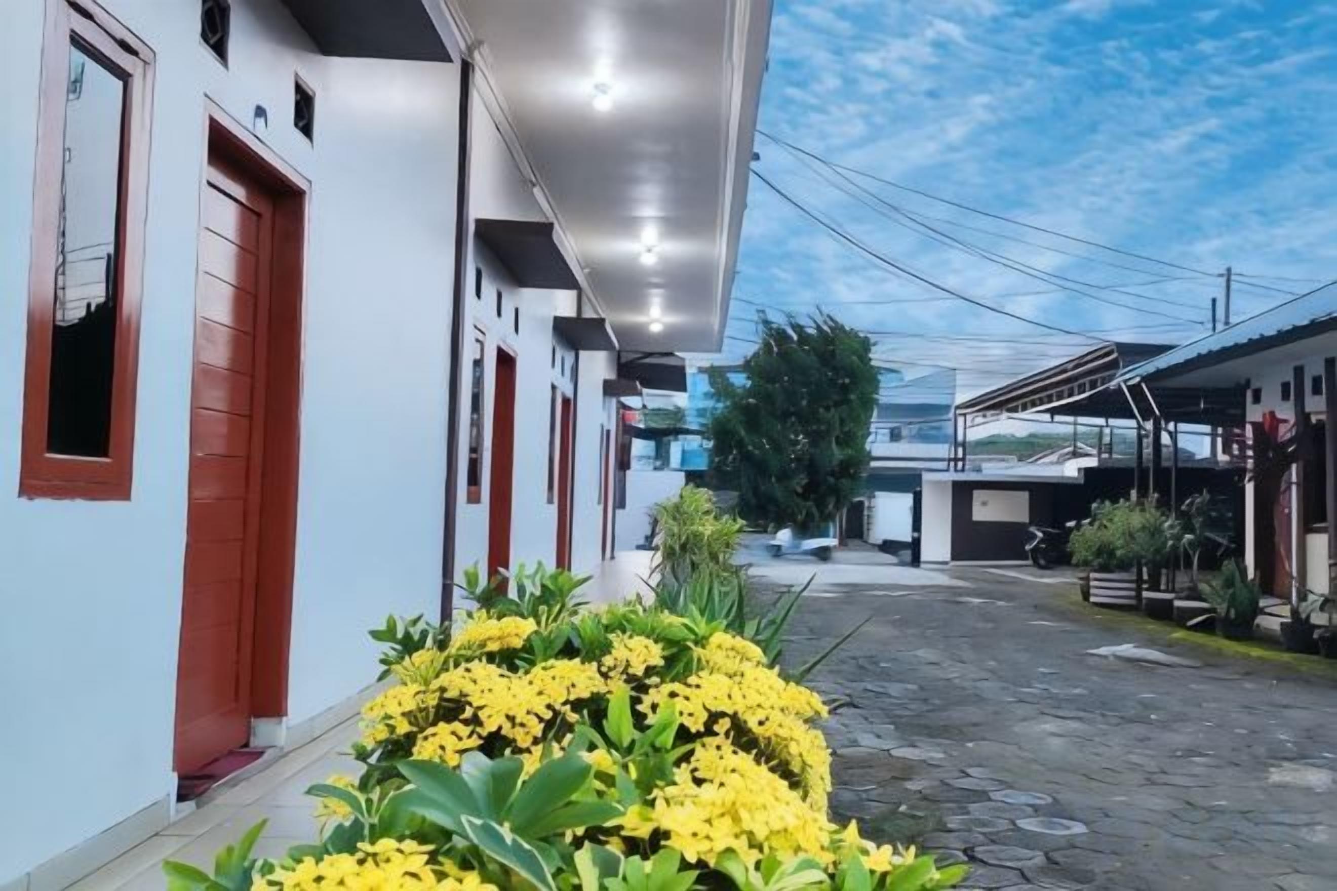 A narrow street with buildings on either side, a clear sky, and a small garden with yellow flowers.