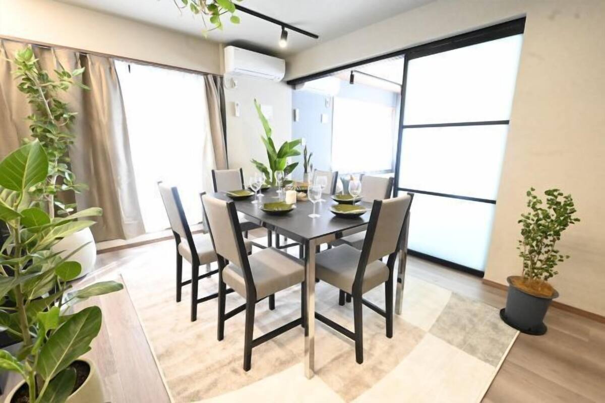 A dining area with a table set for four, surrounded by chairs, with large windows and potted plants.