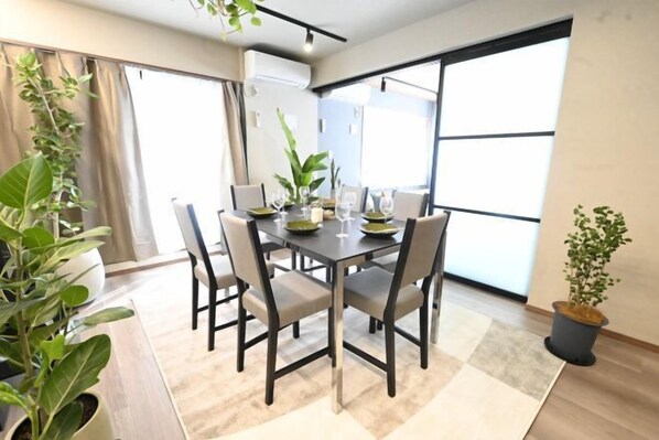 A dining area with a table set for four, surrounded by chairs, with large windows and potted plants.