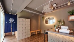 A reception area with a wooden floor, a counter with a white sofa, a mirror, and a plant. There are lockers in the background. - NiHITARU Stay (Tokyo)