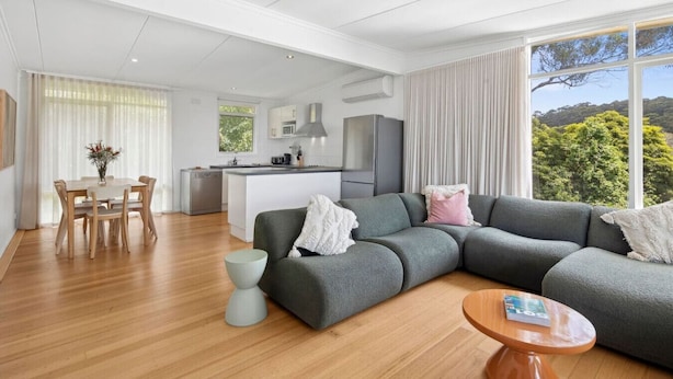 A modern living room with a grey sectional sofa, a dining table with chairs, a kitchen area, and large windows offering a view of greenery. - Prasino in Central Lorne (Lorne)