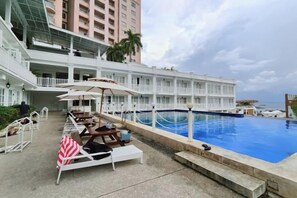 Un espace piscine d’hôtel avec des chaises longues et des parasols, une grande piscine et un bâtiment doté de balcons.