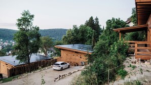 Una baita di legno con un'auto parcheggiata davanti, circondata da alberi e vista su un villaggio sullo sfondo.