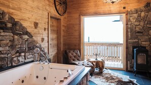 Interno di una baita in legno con parete in pietra, una vasca da bagno grande, un camino e un balcone con vista.