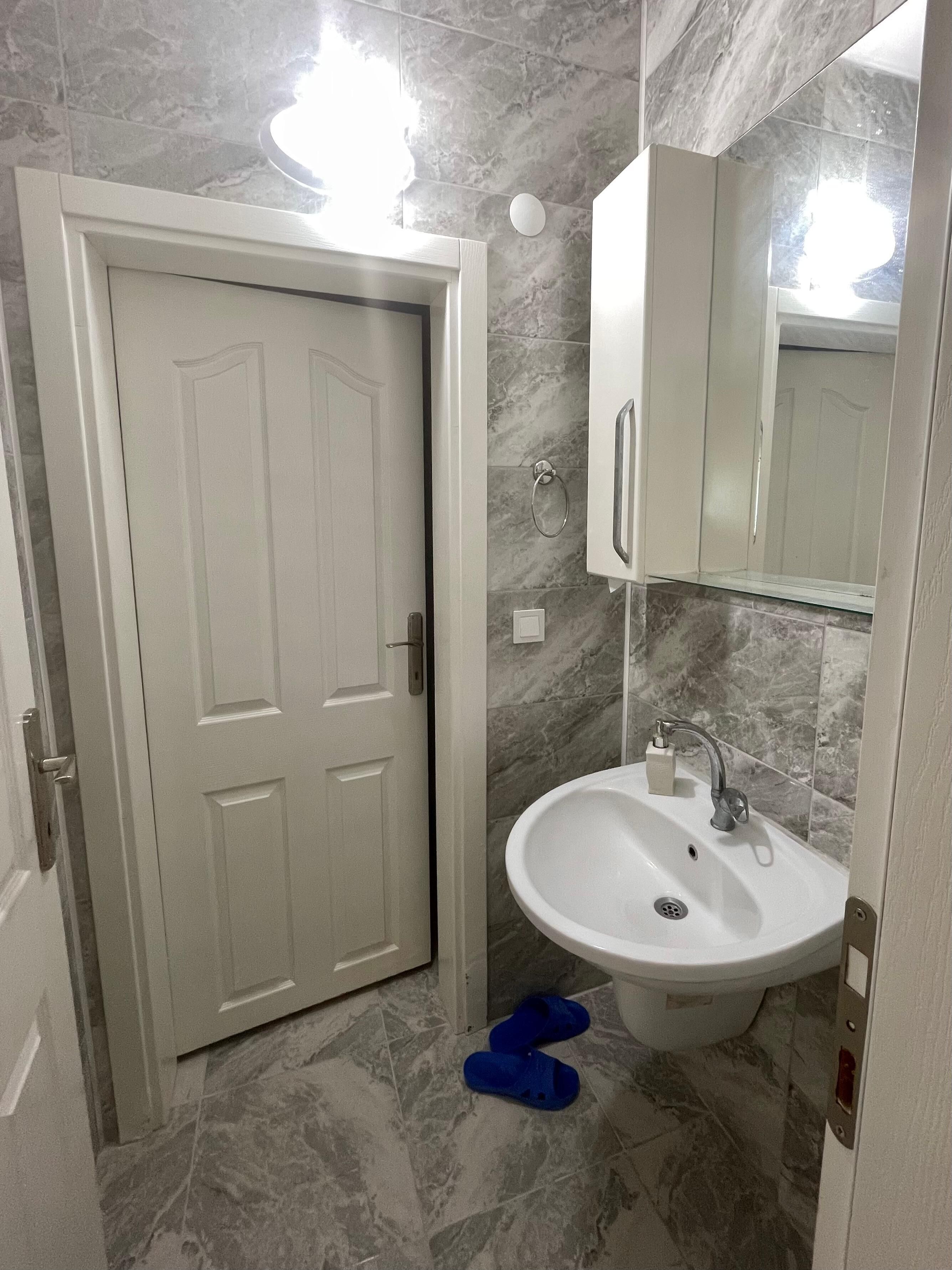 A bathroom with a white door, a sink with a mirror above it, and a pair of blue slippers on the floor.