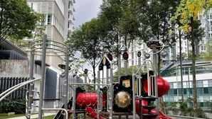 A playground with various play structures, including a red slide and climbing frames, surrounded by trees and buildings.