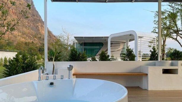 A modern outdoor hot tub area with a wooden deck, a white structure with a slanted roof, and a mountain backdrop. - 101 Space Station (Lan Sak)