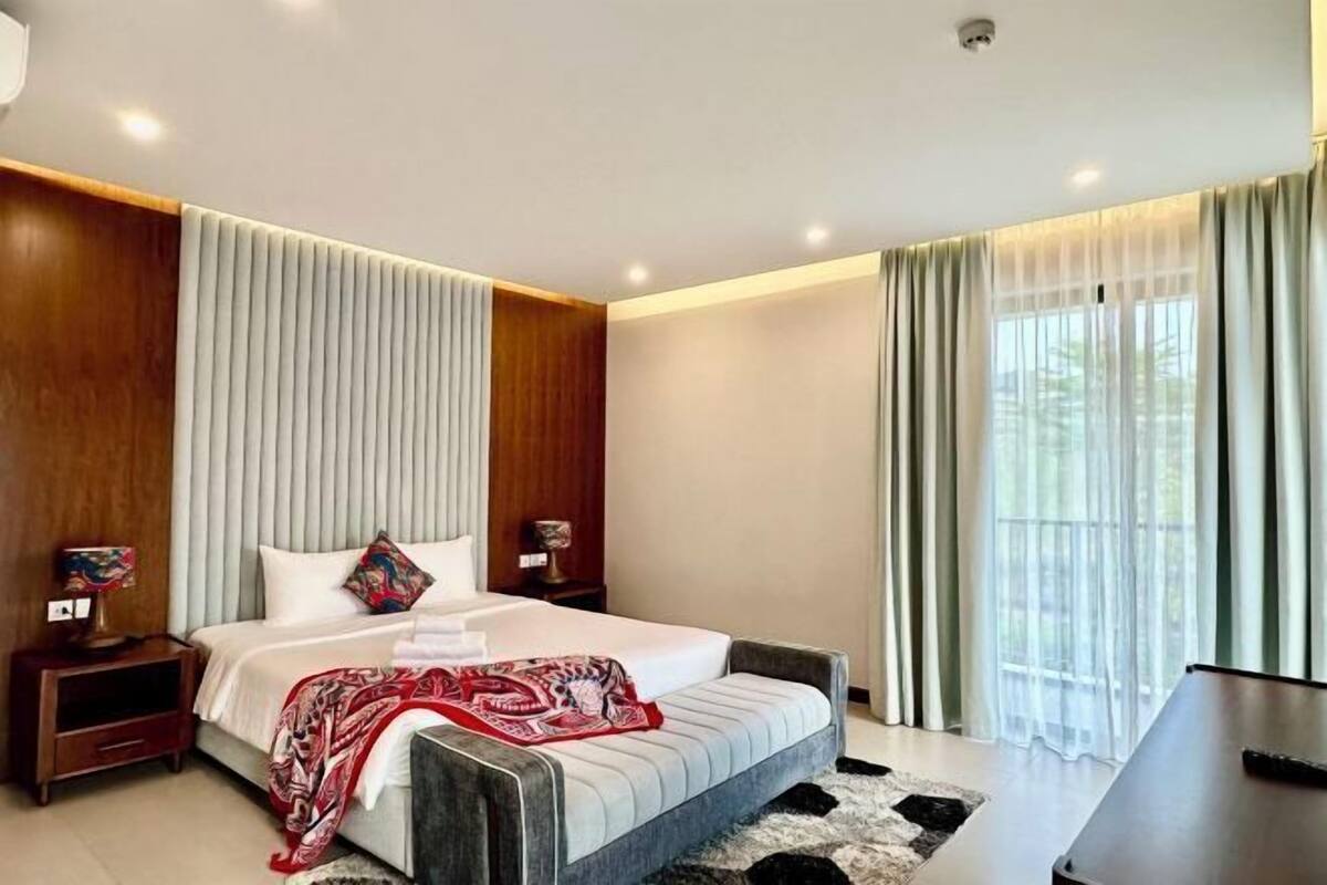 A modern bedroom with a large bed, wooden paneling, a nightstand with a lamp, and a view of greenery through the window.