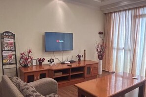 Una sala de estar con televisor de pantalla plana montado en la pared, mueble de madera con objetos decorativos, un sofá y un ventanal con cortinas.
