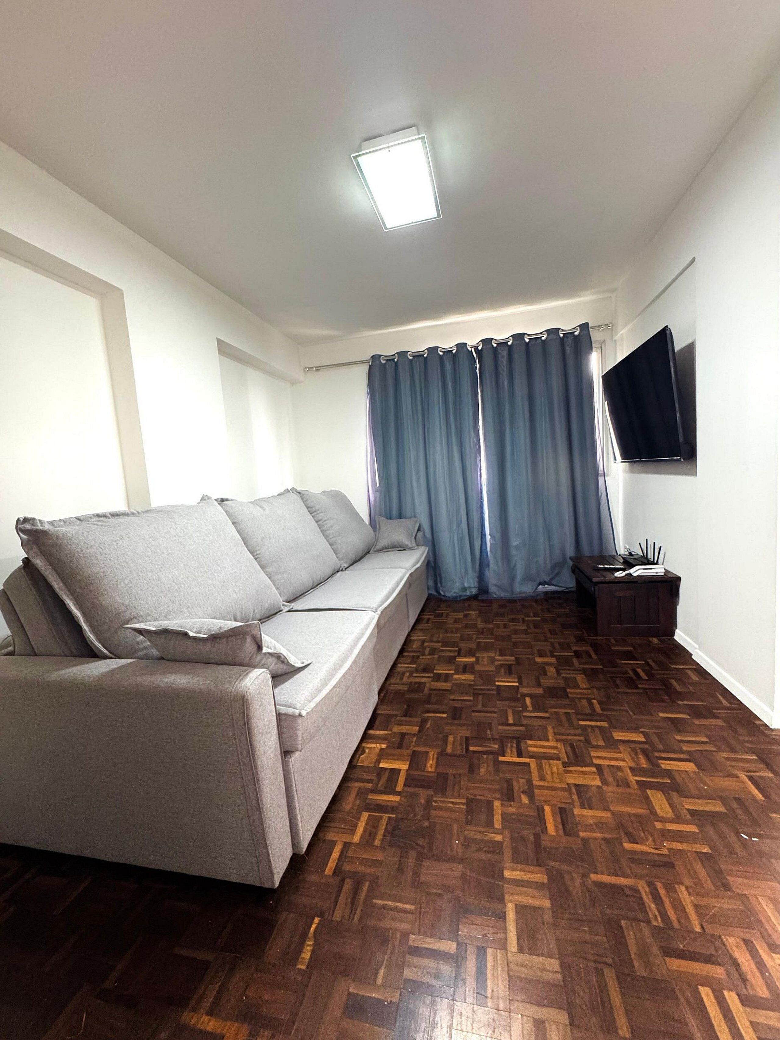 Una sala de estar con sofá, suelo de parqué, televisor en la pared y cortina.