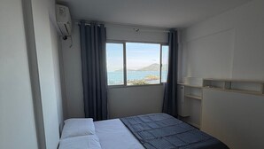 A room with a bed, a window with curtains, a radiator, and a view of the sea. - Edificil Antares (Balneário Camboriú)