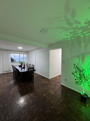 Uma área de refeições com piso de madeira em padrão herringbone, uma mesa com cadeiras, uma janela com vista e uma planta verde na parede.