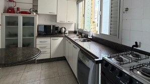 A compact kitchen with white cabinets, a dishwasher, a stove, and a window providing natural light. - Edificil Antares (Balneário Camboriú)