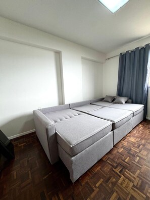 Quarto com sofá-cama, janela com cortinas e piso em assoalho de madeira.