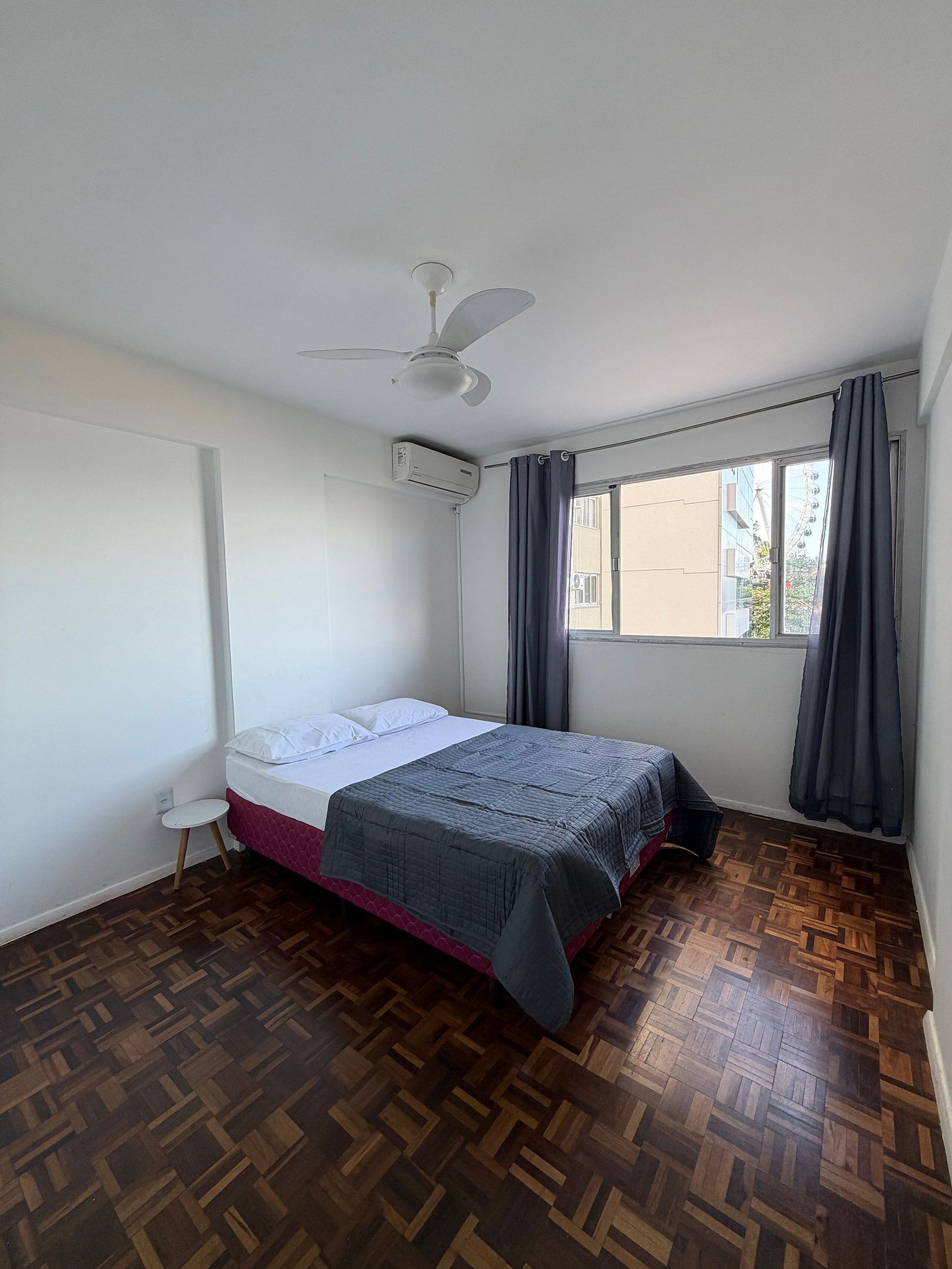 A bedroom with a bed, a ceiling fan, an air conditioning unit, a window with curtains, and a wooden parquet floor.