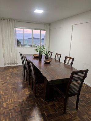A dining room with a large wooden table, six chairs, a potted plant, and a view of the sea through a large window. - Edificil Antares (Balneário Camboriú)