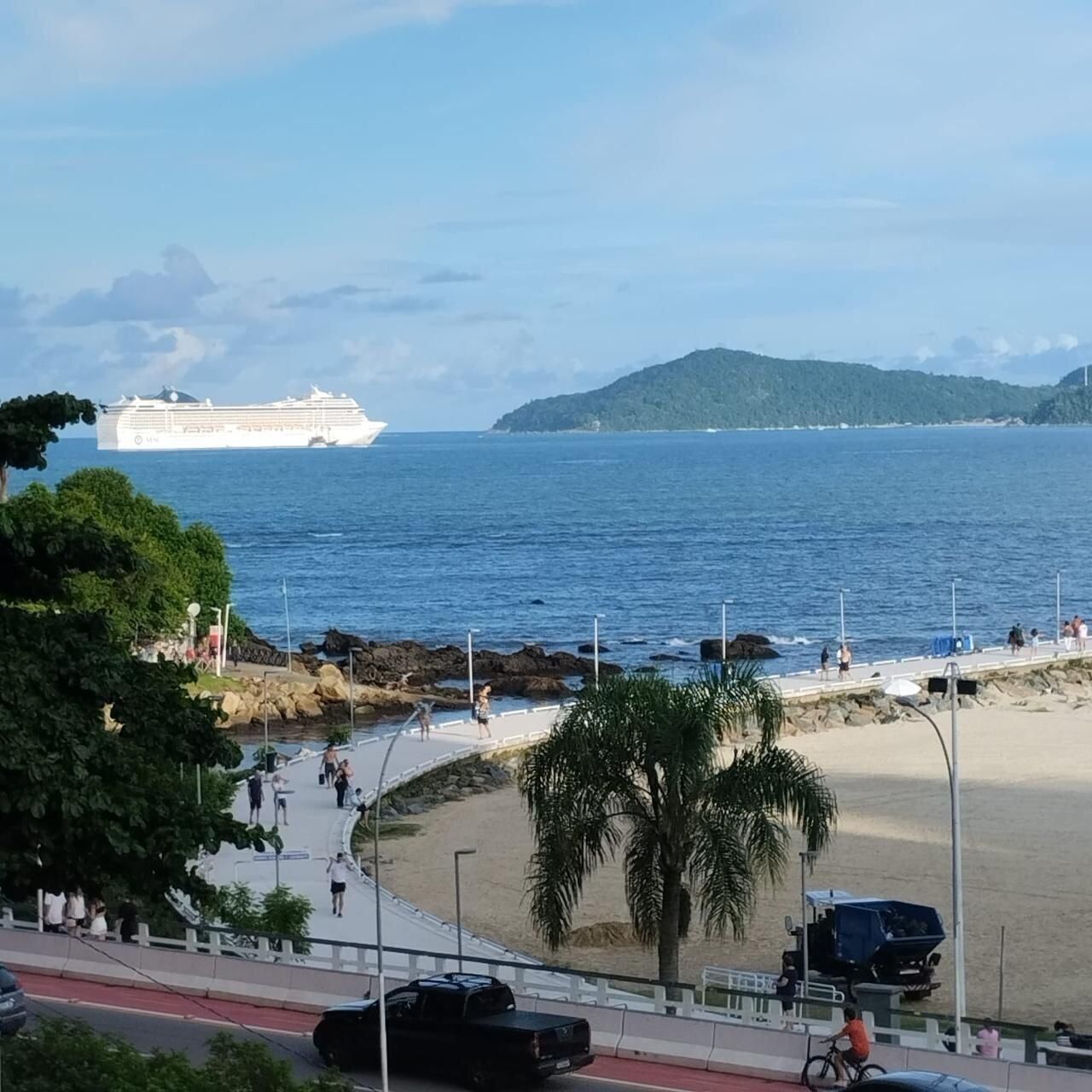 Cena costeira com um cruzeiro, praia, palmeiras e uma estrada com veículos.