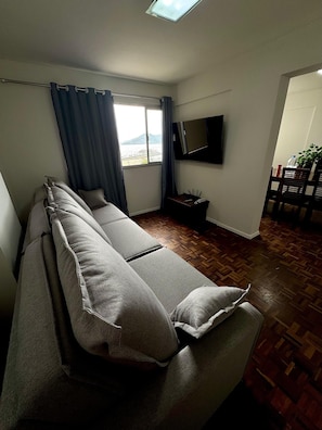 A room with a sofa, a TV, a window with curtains, a wooden floor, and a dining area with chairs.