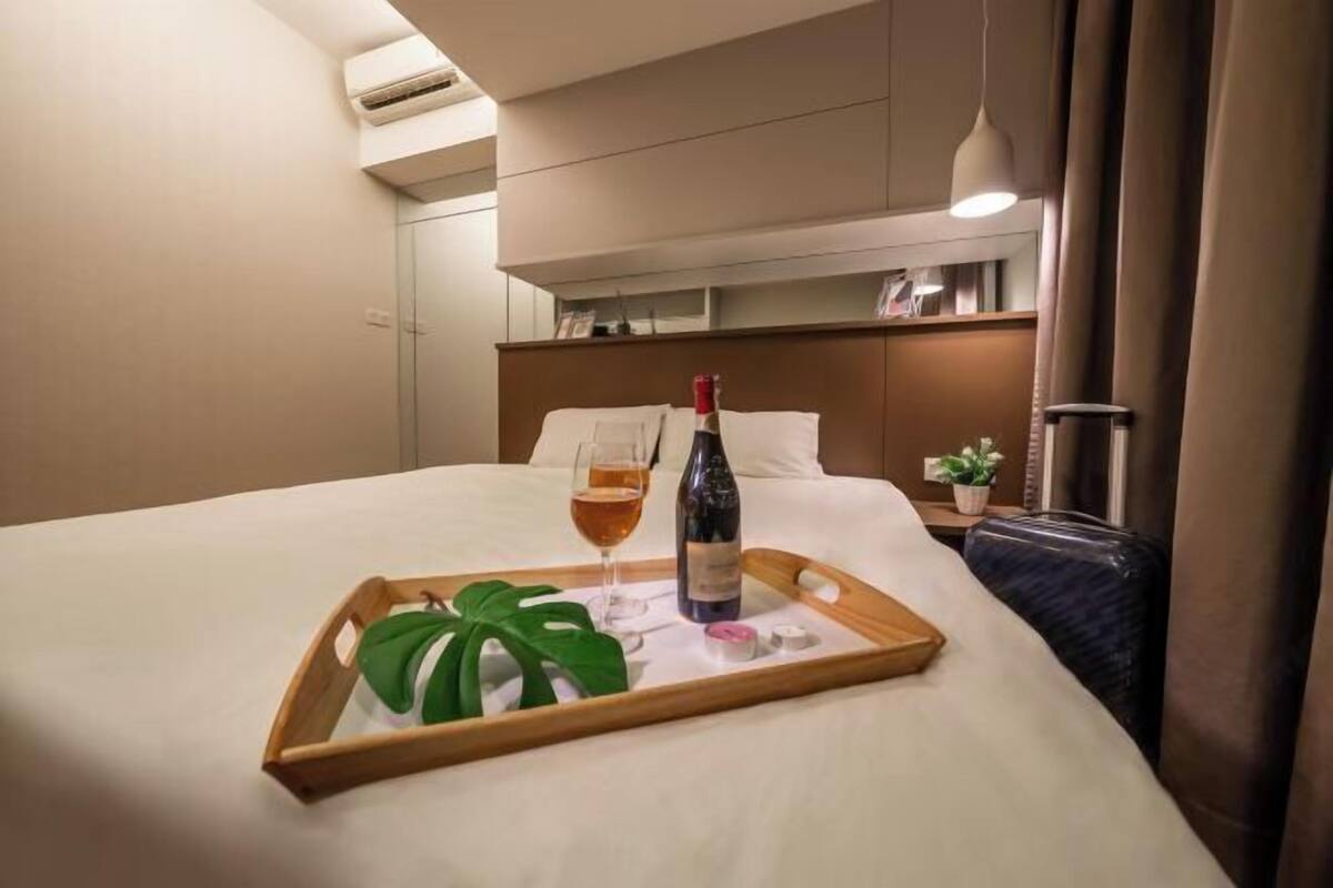 A hotel room with a bed, a bedside table with a bottle of wine and a glass, a tray with a plant, and a suitcase.