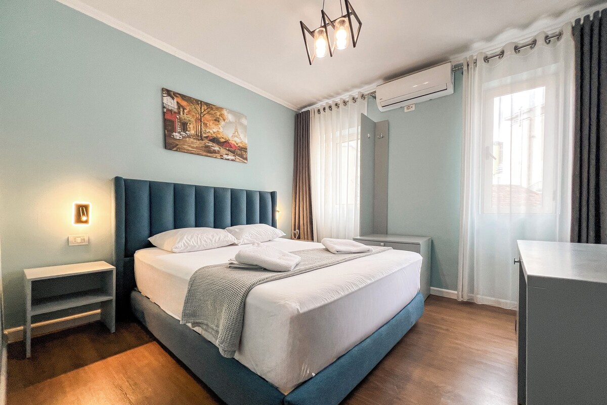 A neatly arranged bedroom with a large bed, bedside tables, a nightstand, a desk, and a painting on the wall.