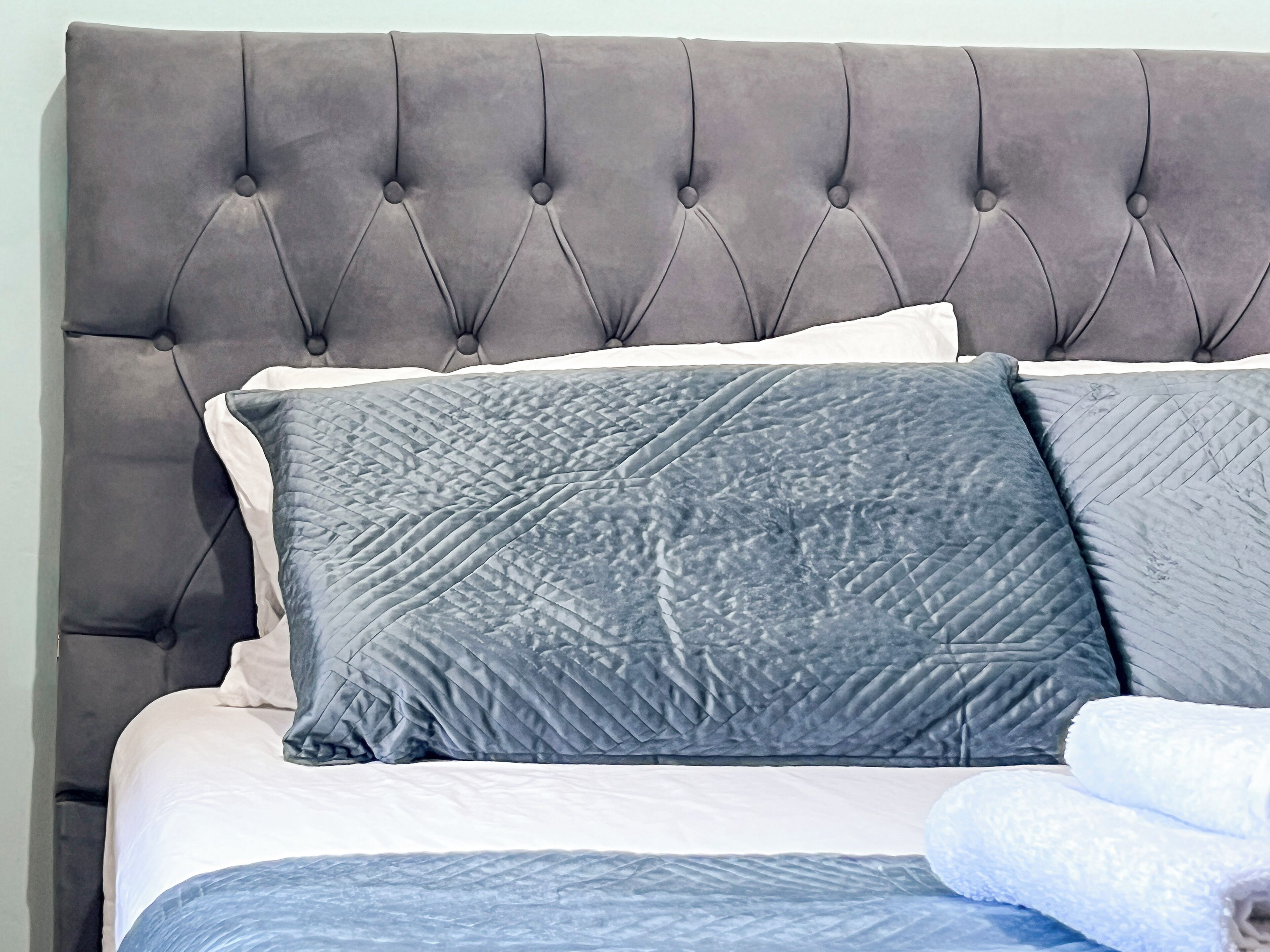 A bed with a tufted headboard, grey bedding, and a folded white towel.