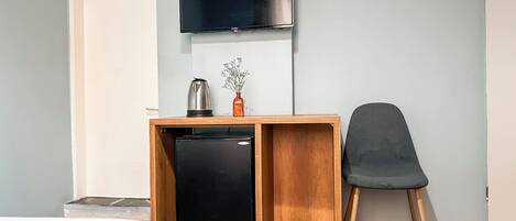 Una habitación compacta con un escritorio de madera, un refrigerador negro, una silla, un televisor fijado a la pared y un pequeño florero con flores.