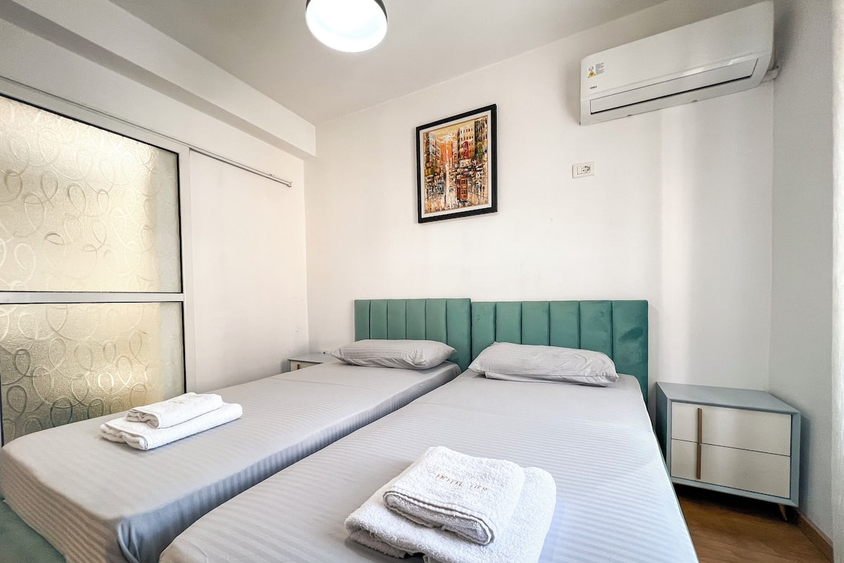 A hotel room with two single beds, a nightstand, an air conditioning unit, and a framed cityscape painting.