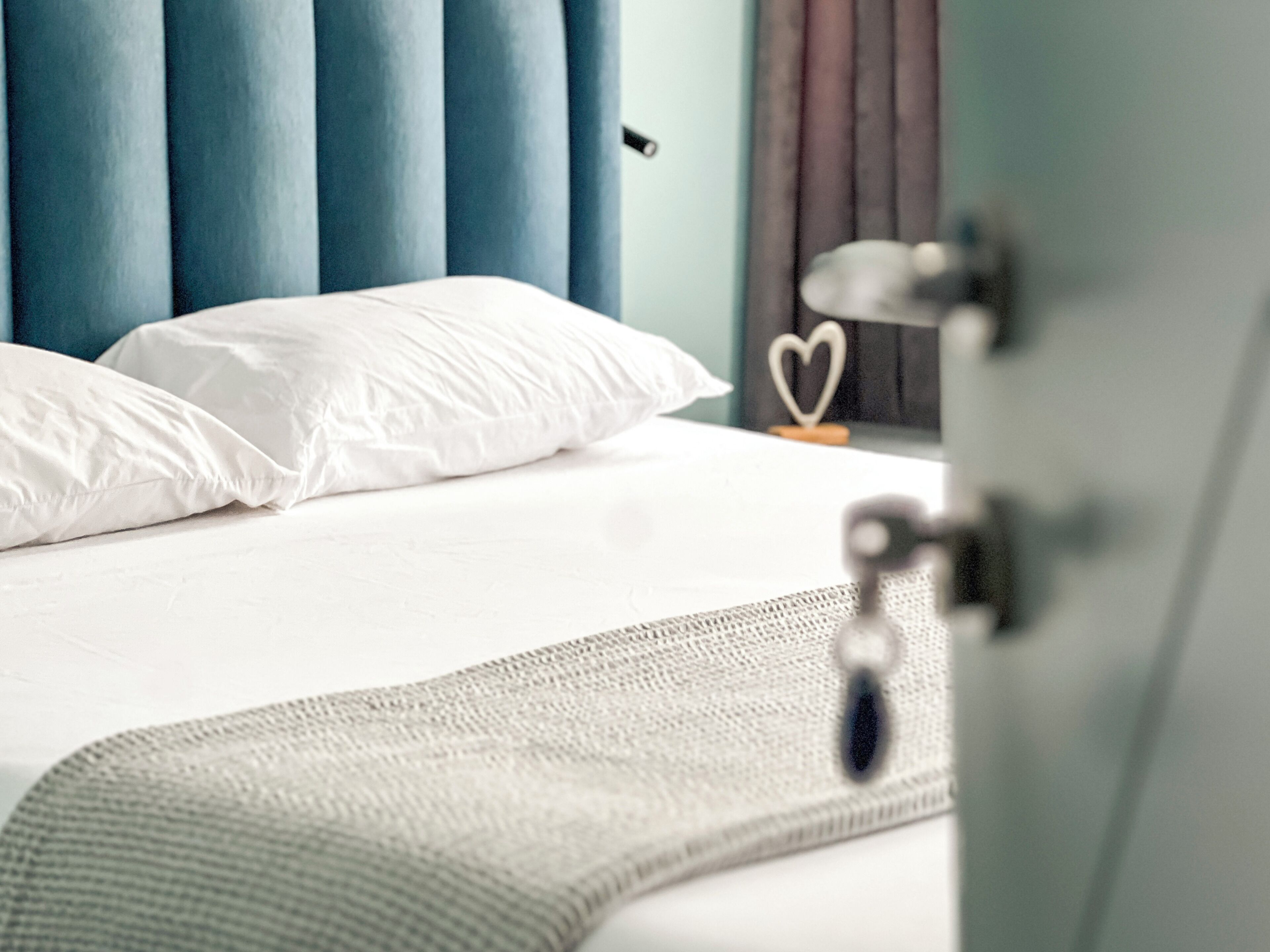 A neatly made bed with white linens, a blue headboard, and a heart-shaped decoration on the wall.