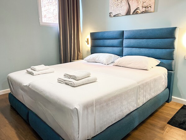 Un lit bien fait, avec du linge de lit blanc et des serviettes pliées, une tête de lit bleue en tissu et un tableau représentant des cerfs accroché au mur.