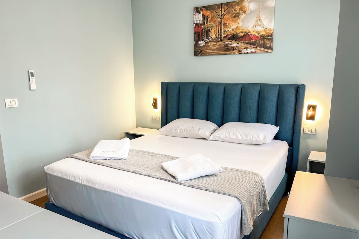 A neatly made bed with white linens and a blue headboard, a painting of a Parisian street scene, and a small bedside table with a lamp.