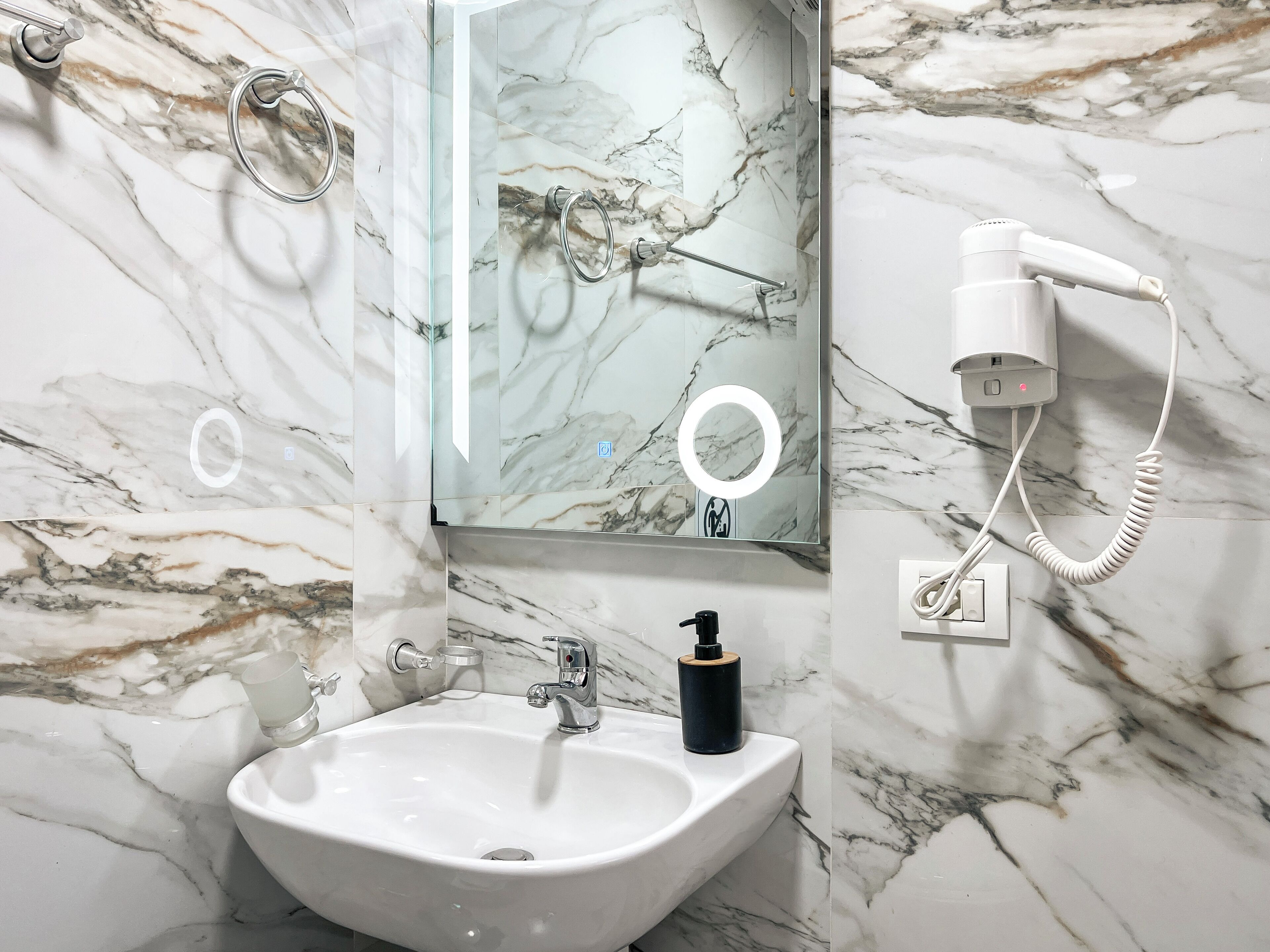 A bathroom with a marble sink, a mirror, a hairdryer, and a soap dispenser.
